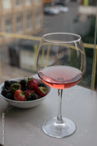 Obraz Kieliszek wina różanego znajduje się na stole na balkonie, obok talerza jagód. Pij wino i jedz jagody. Nastrój lata. Wino z owocami. Letnie wakacje z winem. Napoje alkoholowe.