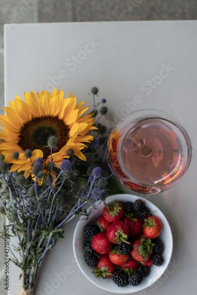 Obraz A glass of rose wine stands on the table on the balcony, next to a plate of strawberries and blackberries and a bouquet of flowers made of sunflower and blue thorn. Drink wine. Summer day off. 