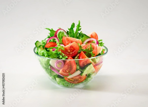 Fototapeta Healthy food. Vegetable salad in a transparent bowl. Salad of red tomatoes, cucumbers, red onions, green lettuce leaves, garlic with olive oil on a white background. Background image, copy space