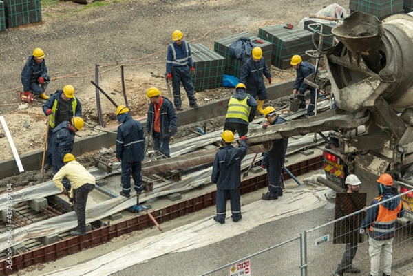 Fototapeta Tel Aviv, Israel - May 20 2021: Construction Workers working in the rain. Light rail tracks. blue collar worker. Concept Collaboration teamwork. Trucks, concrete mixer, bulldozer. High quality photo