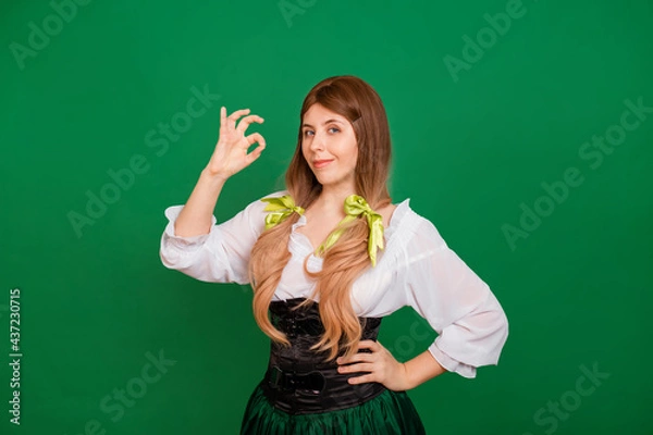 Fototapeta Young woman in clothes for celebrating St. Patrick's Day and Oktoberfest showing okay gesture and smiling isolated on green background.