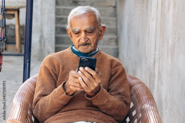 Obraz Happy elderly grey-haired Indian man in casual brown clothes using mobile phone, typing sms message. Old people lifestyle concept	
