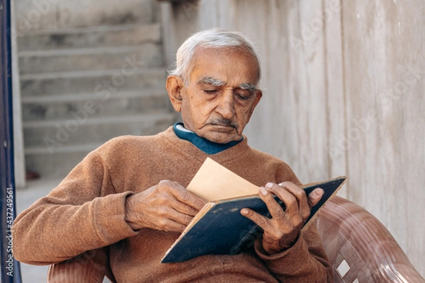 Obraz Portrait of old Indian man turning pages of old diary while sitting on chair at home. Leisure activity of retired people. Senior people lifestyle	
