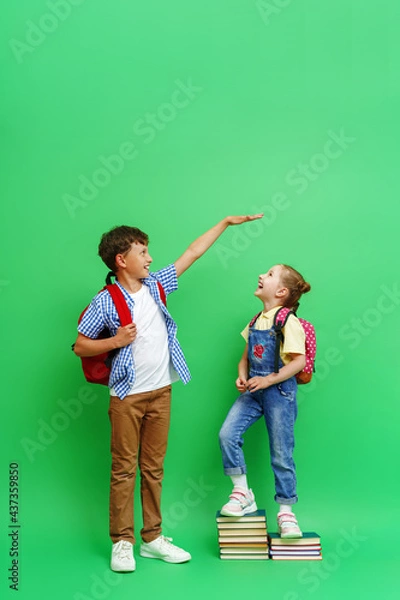 Fototapeta happy teenage schoolboy with a backpack, measuring the height of a little girl in loose clothes, standing on a stack of books on a green background. The concept of development and growth of education.