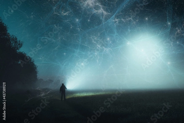 Fototapeta A science fiction concept of a man standing in a spooky field in the countryside, holding a torch. With apocalyptic clouds of energy glowing in the sky.