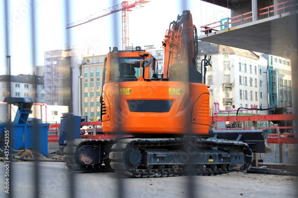 Fototapeta Orange crawler excavator on a construction site in the city centre behind a construction fence.