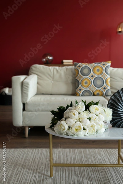 Obraz bouquets of white peonies lie on the coffee table. In the background is a white sofa with a colorful pillow. The living room has red walls. A bouquet of white flowers. Fresh flowers in the interior.