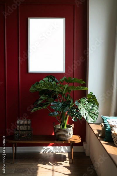 Obraz A monstera flower stands in a wicker basket on a coffee table against a red wall. A white frame hangs on the wall. A place for a picture or poster. Modern interior. Sunlight shines on the wall.