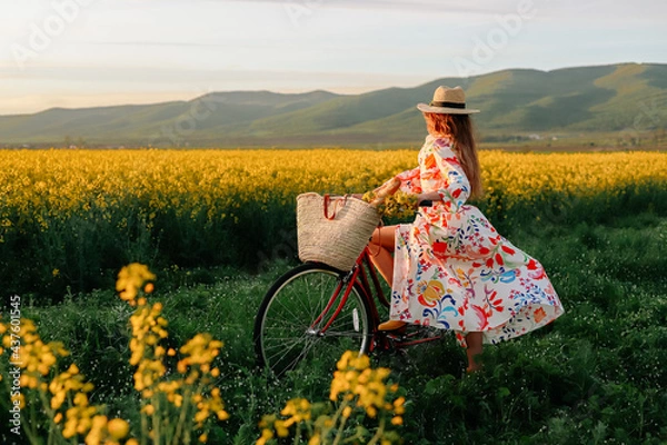 Fototapeta Girl with a red retro bicycle going along the yellow field of flowers.