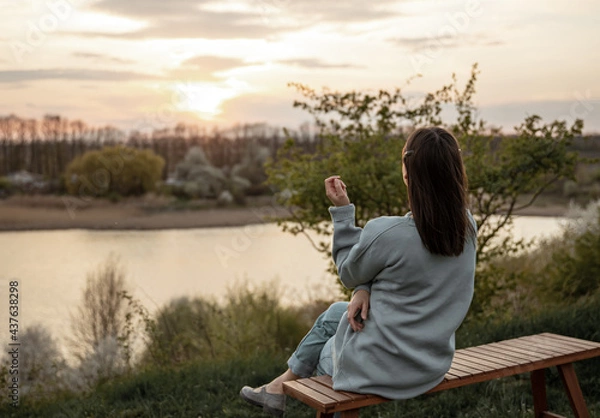 Fototapeta A girl in casual style sits and looks at the sunset near the lake.
