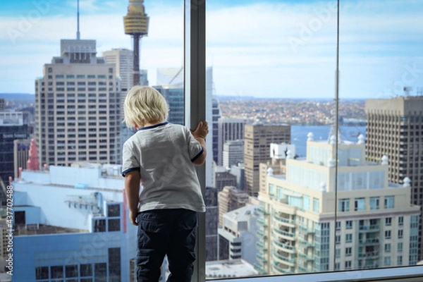 Fototapeta Little boy standing at window of high rise apartment looking down at view of the city below