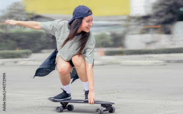Fototapeta Beautiful happy Asian healthy woman smiling, motion speed riding and playing extreme sportive skateboard as outdoor activity with happiness, relaxation and fun during holidays in summer vacation.
