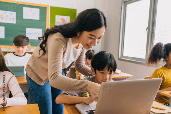 Obraz Young adult smiling beautiful Asian teacher helping elementary student boy with laptop in computer classroom. Information Technology class in primary school concept.