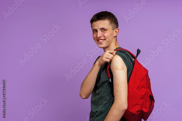 Fototapeta Happy Smiling Vaccinated Young Man Student, With Backpack Showing His Hand With Patch After Injection Covid-19 Vaccine, Poses On Purple Background. Advertising of vaccination against coronavirus.