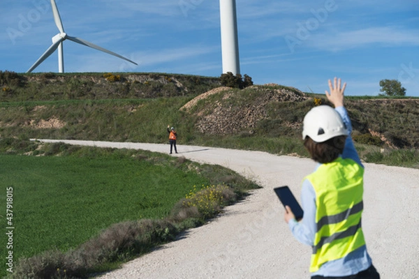 Obraz Focus on the worker in the orange vest. Unrecognized engineers wearing safety clothes working in wind turbine farm. They are making tests.