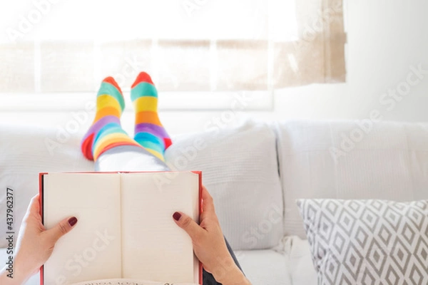Fototapeta Lesbian woman relaxing at home on a sofa reading a book and dressed in rainbow colored socks. LGBT Concept