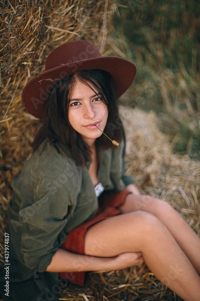 Fototapeta Beautiful stylish woman with herb in mouth and in hat sitting at haystack in summer evening field. Portrait of sexy young female relaxing at hay bale, summer vacation in countryside.