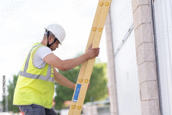 Fototapeta telecommunications technician coming down a yellow ladder and looking down, wearing a helmet and reflective vest.