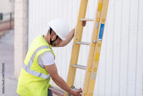 Fototapeta telecommunications technician with safety helmet and reflective vest, deploying a yellow ladder while looking down.