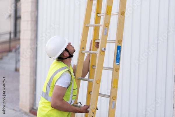 Fototapeta telecommunications technician in safety helmet and reflective vest, deploying a yellow ladder while looking upward.