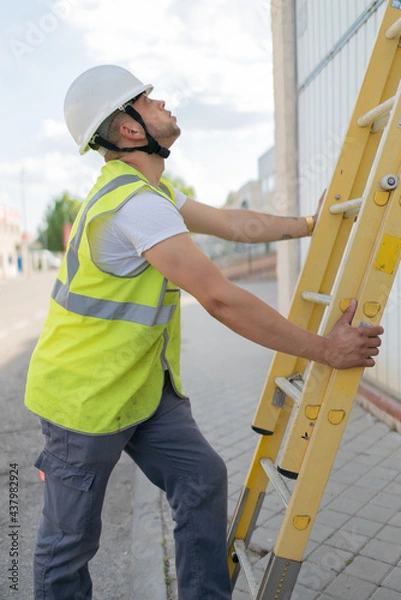 Fototapeta telecommunications technician holding a ladder looking upward, wearing a safety helmet and reflective vest