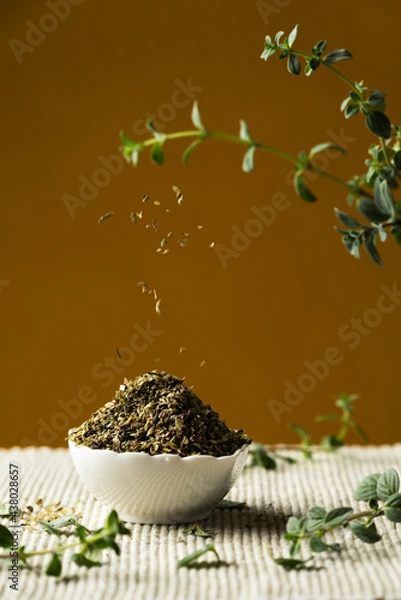 Fototapeta Heap of dried oregano in a white porcelain bowl, and shredded oregano falling, with fresh twigs and leaves, on a yellow or mustard color background, vertical image.