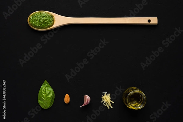Fototapeta Top down view of basil pesto sauce in a wooden spoon and a basil leaf, almond, garlic, parmesan cheese and olive oil as ingredients on a black surface with copy space