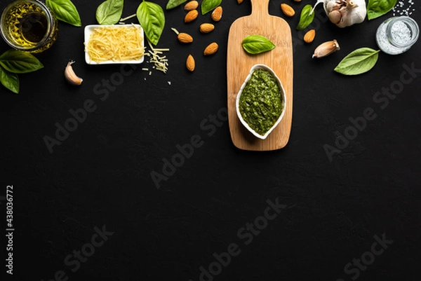Fototapeta Pesto sauce in a white leaf-shaped bowl and a little wooden table with basil leaves, parmesan cheese, almonds, garlic, olive oil and salt as ingredients on a black surface with copy space