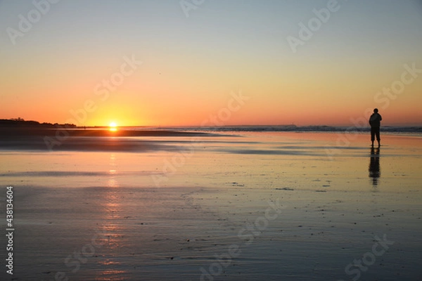 Fototapeta Man watching the sunrise on the beach, with warm tones and a beautiful reflection of the sunlight in the water