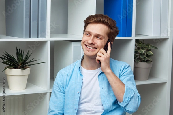 Fototapeta Young attractive man speaking on smartphone while standing in office