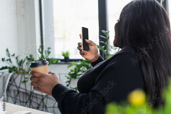 Fototapeta african american plus size businesswoman looking at smartphone with blank screen while holding paper cup.