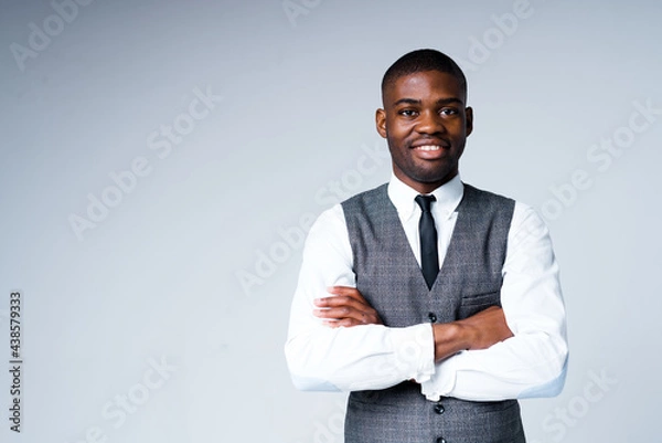 Obraz Handsome african american man in a shirt and vest stands on a light background with his arms crossed on his chest