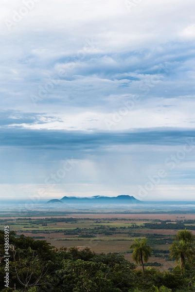 Fototapeta Beautiful view of the hills and open field in Paraguari, Paraguay, with the rain falling over the hills on the horizon and layers of clouds above them, trees in the foreground.
