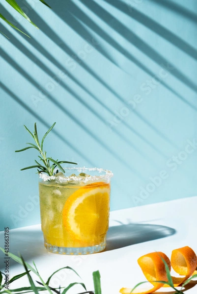 Fototapeta Iced orange refreshment juice with a slice inside the glass, rosemary as a garnish and a lot of ice, on a cyan background with palm tree shadows and space for text