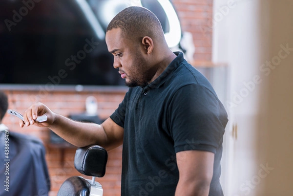 Fototapeta young barber in latin barber shop. with razor in hand to work. resting his arm on the barber's chair. with led light behind the head..