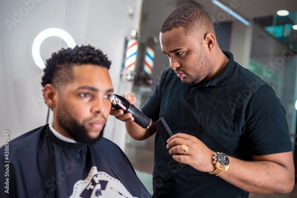 Fototapeta hairdresser cutting the hair of a young man in a latin barber shop.with haircutting machine carefully in front of the mirror.with watch and ring.and led light ring behind the client..
