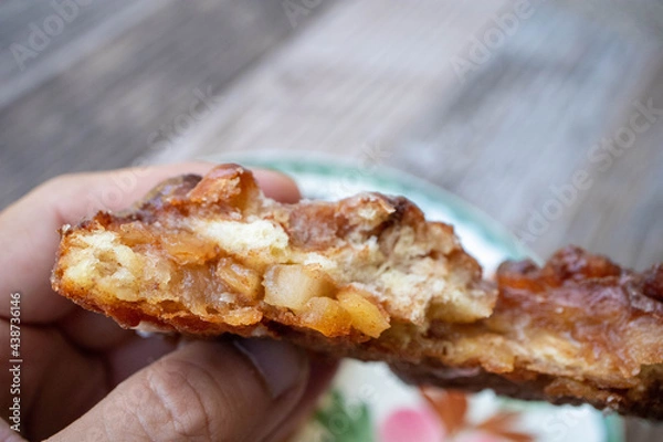 Fototapeta A hand holding n apple fritter pastry showing pieces of apples and cinnamon on the inside over a plate on a table