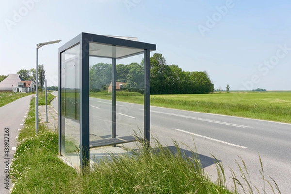 Fototapeta Bus stop shelter with glass on a road in a rural area under a blue sky.