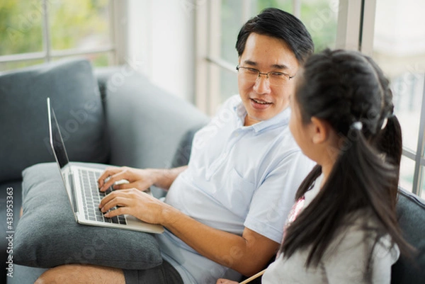 Fototapeta Young Asian love family father and daughter teach homework by computer notebook on sofa in living room at home which smiling and felling happy. live at home concept in COVID-19 situation