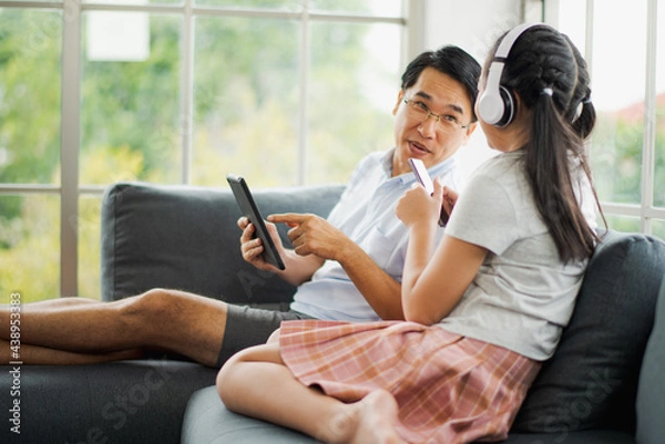 Fototapeta Young Asian love family father and daughter use tablet and smart phone to listen music on sofa in living room at home which smiling and felling happy. live at home concept in COVID-19 situation