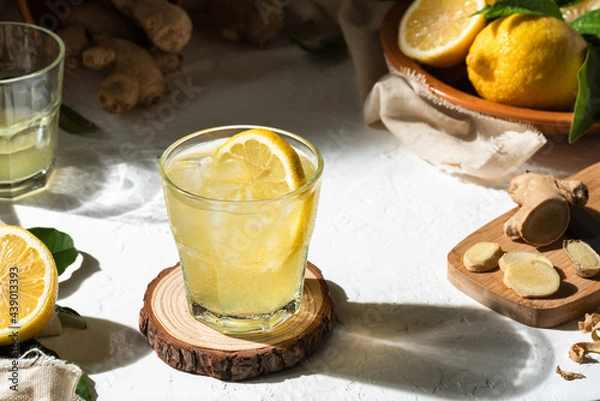 Fototapeta Detox lemon and ginger drink, with ice and a slice of lemon inside the glass, as an immune boosting and weight loss drink on a white textured surface, hard light