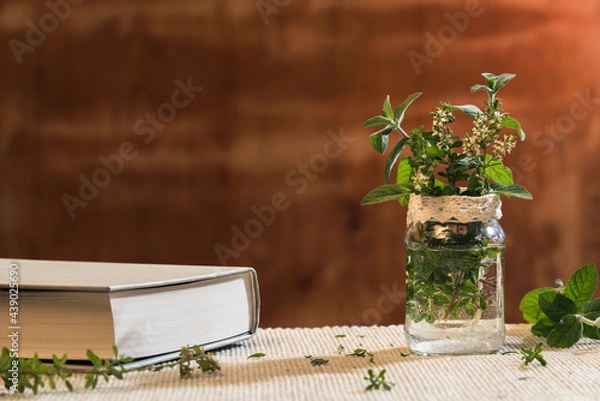 Fototapeta Fresh twigs of thyme and peppermint in a glass jar with water on a natural morning light, and a book in the left third. Wooden background with space for text. Aromatherapy and reading concept.