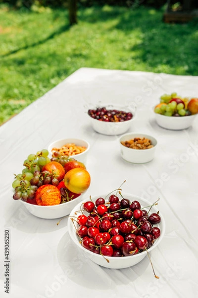 Fototapeta stylish vertical food content. ripe cherry berry, ripe juicy apples, grapes and nuts in white paper plates on light table in an open space green grass. healthy summer raw snacks. selective focus