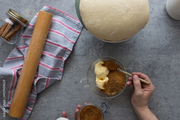 Obraz Woman hand pouring cinnamon powder with a spoon in a bowl full of panela and butter next to a risen dough.