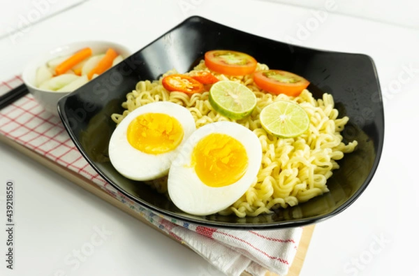 Fototapeta Indonesian noodle With a black bowl with eggs and fresh tomatoes on a napkin base on a white background
