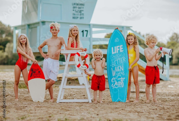 Obraz Kids posing on the sand beach with lifeguard tower and surfboard. Happiness.