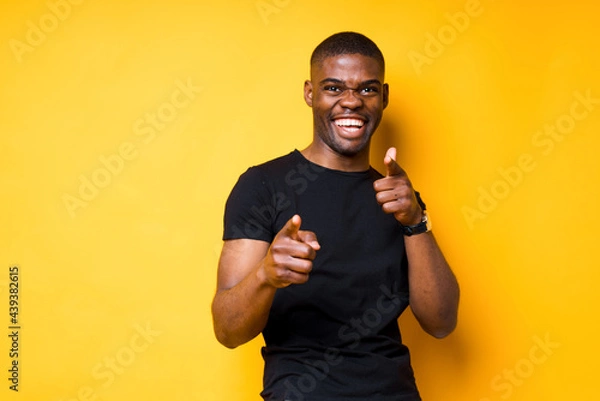Obraz Cheerful african american man in black t-shirt smiles and points fingers at camera