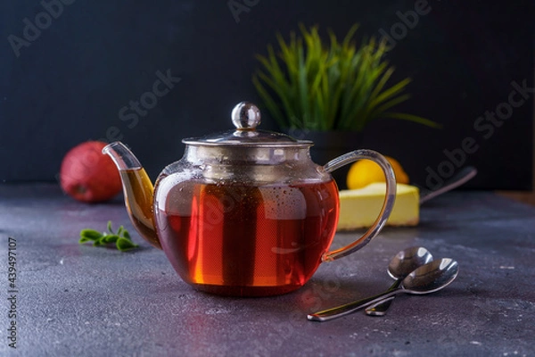 Obraz Black tea in a clear glass teapot on a dark background. There's a cake in the back