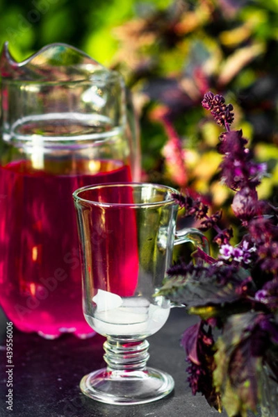 Fototapeta Glass with ice on a dark table with decanter full of purple basil coctail background. Picnic in thу summer garden with trees and leaves.