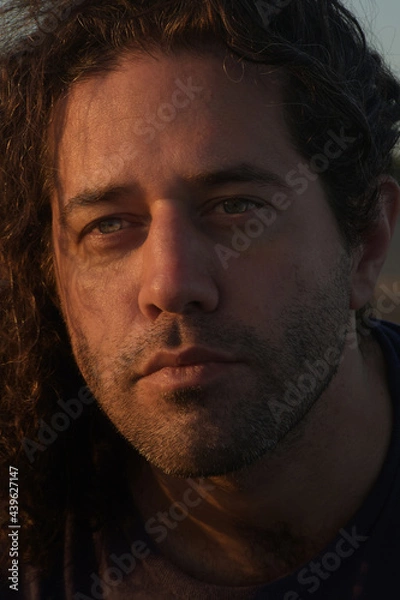 Fototapeta Handsome man with light eyes and deep gaze looking forward with his curly hair. Isolated portrait with warm tones and soft sunlight on the face